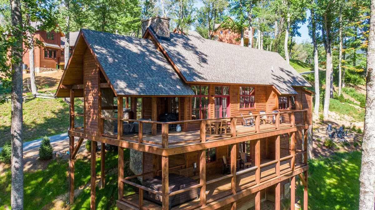 A wide wooden deck stretches along the exterior of the cabin, framed by tall trees and anchored by a striking stone chimney. Large windows offer a glimpse of the interior, while the deck itself is furnished for outdoor relaxation and dining. The scene feels peaceful and invites group gatherings with mountain air and scenic forest views.