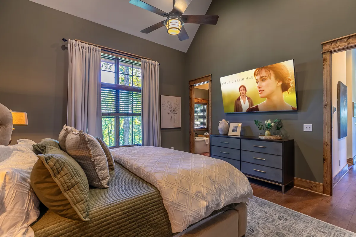 A luxurious King Suite bedroom featuring a plush bed with decorative pillows, a window letting in natural light, a sleek wall-mounted television displaying a film scene, and a nightstand. The setting feels both restful and upscale, offering space to unwind and enjoy private entertainment.