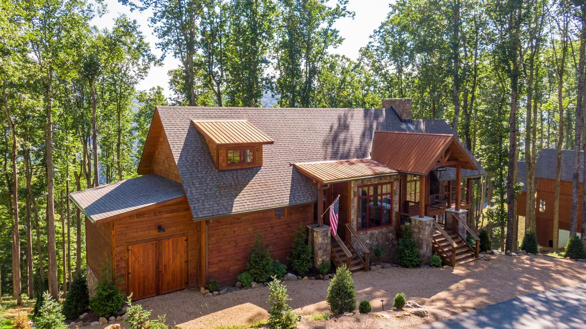 The image showcases the inviting wooden exterior of a newly built mountain home nestled among towering trees. Large windows reflect the surrounding nature, a welcoming front porch is draped with an American flag, and the scene exudes warmth and tranquility—perfect for gatherings or relaxation. The natural setting hints at sunset views and peaceful mornings enjoying coffee outdoors.