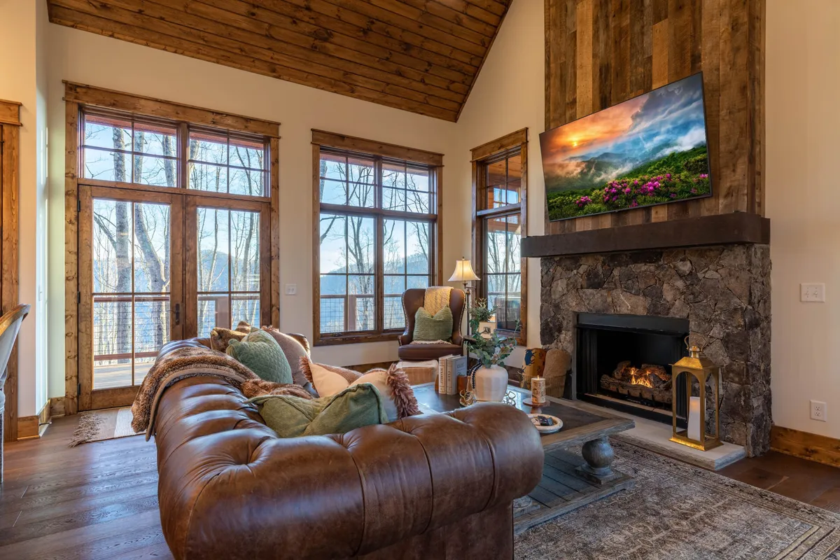 A cozy living room with a stone fireplace as the centerpiece, plush leather couch, large windows revealing a forest view, and a television displaying a scenic image. The arrangement invites relaxation, socializing, and enjoying entertainment.
