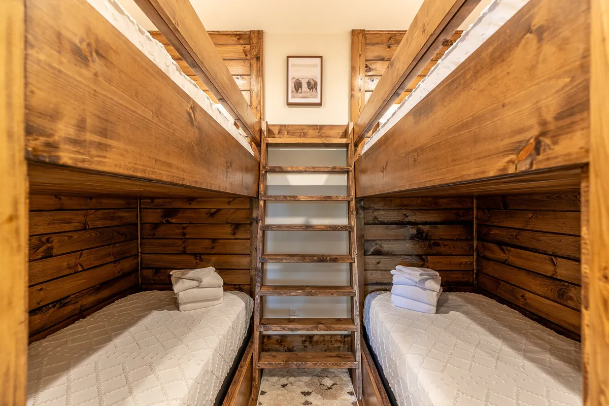 A playful and inviting bunk room, with two sets of bunk beds arranged to maximize sleeping space. A wooden ladder provides access to the upper bunks, and a framed picture adds a touch of personality. The room feels cozy and functional, ideal for children or extra guests.
