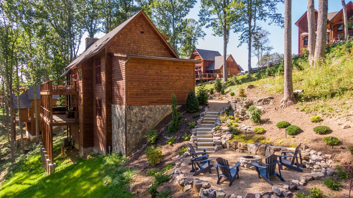 A cozy outdoor area featuring a wooden cabin, a stone fire pit, comfortable seating, and lush greenery. The space suggests opportunities for gathering, storytelling, and enjoying the outdoors, especially in the evenings.