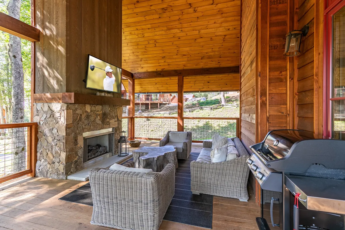 An inviting outdoor living area features a stone fireplace, a mounted TV, a grill, and a comfortable seating arrangement. The space is designed for group entertainment—perfect for watching sports, sharing meals, or simply relaxing while surrounded by greenery and open skies.