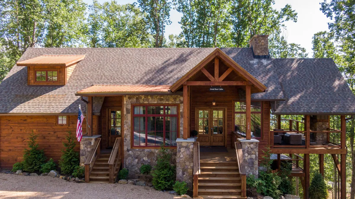 A welcoming exterior shot of a wooden structure with a large front porch, stone facade, American flag, and abundant greenery. The porch is set up for comfortable outdoor gatherings, surrounded by nature, perfect for family barbecues and enjoying sunset views.