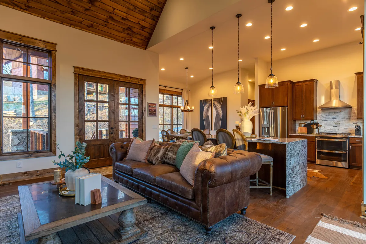 A stylish, open-concept space featuring a leather sofa, kitchen island with pendant lights, large windows that bring in natural light, and a view into the kitchen and dining area. The layout emphasizes social interaction and seamless flow between spaces.