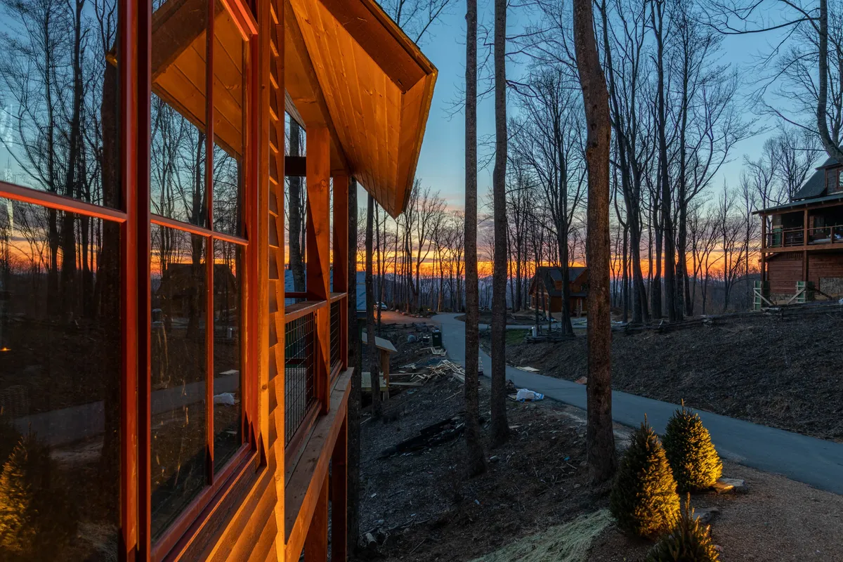 This exterior scene shows a wooden cabin nestled among tall trees, with a glowing sunset in the sky. A gentle pathway winds through the landscape, inviting exploration and quiet evening gatherings under the stars.