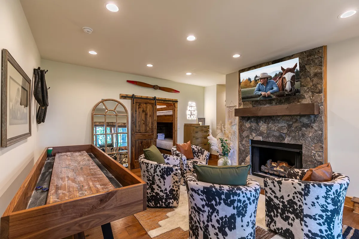 This image features a cozy den with a fireplace, sofa seating, a television displaying a horse scene, and a shuffleboard table. The space is styled for comfort and relaxation, with warm lighting and an inviting ambiance, perfect for family gatherings, movie nights, or game tournaments.