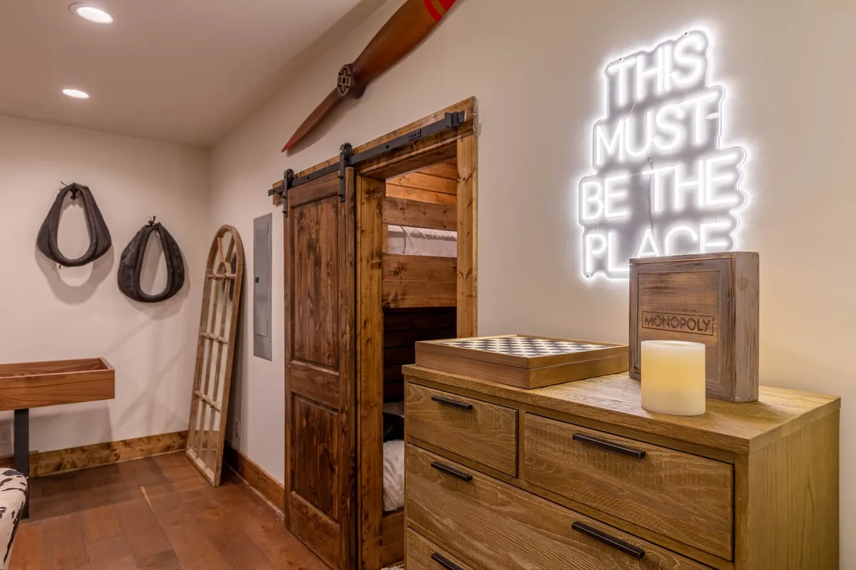 This image shows a cozy interior corner featuring a neon sign, wooden door, chest of drawers, and a checkers board set up for play. The setup invites guests to gather for board games and enjoy a sense of camaraderie and fun.