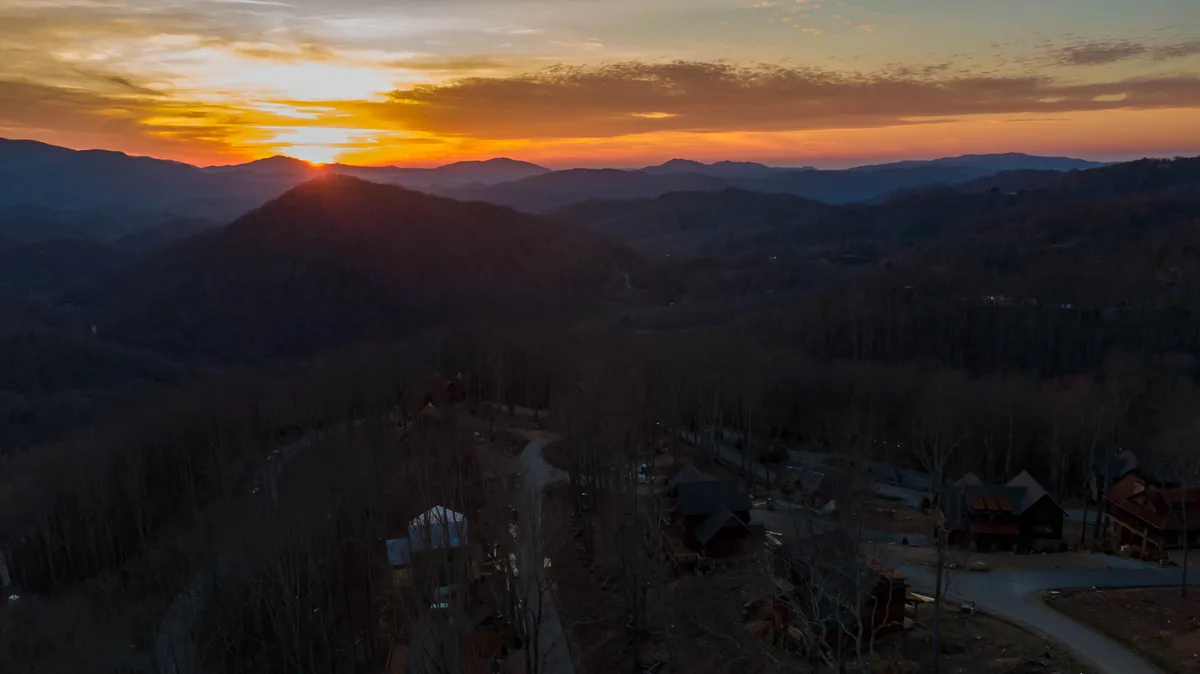A sweeping view of a sunset over the mountains, with a curved road winding through a forest and scattered cabins nestled in the valley. The scene evokes adventure, exploration, and the opportunity for evening campfires and wildlife observation.